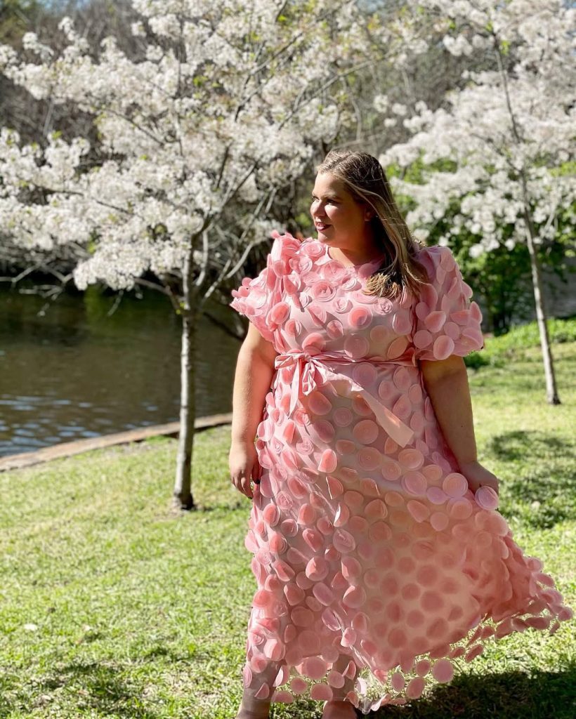 Plus size woman in pink textured dress among cherry blossoms, romantic spring fashion.